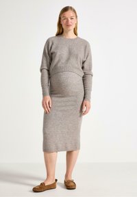 En gravid kvinna står självsäkert, klädd i en grå stickad topp och en figurnära grå klänning, ihop med bruna mockaskor.
