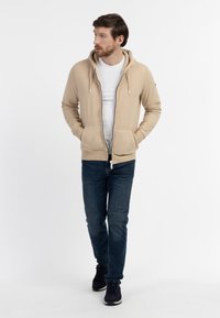 Beige zip-hoodie med snoretræk i huen, forlommer og hvidt indre lag, parret med mørkeblå jeans og marineblå sneakers.