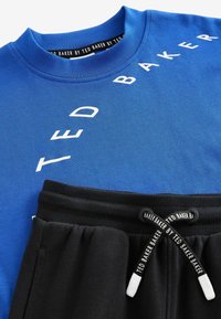 Modrá bavlnená mikina s bielym nápisom "TED BAKER", skombinovaná s čiernymi teplákmi s rebrovaným pásom a šnúrkou na stiahnutie.