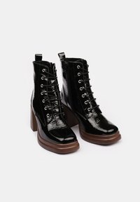 Botas de tobillo de charol negro con un alto tacón cuadrado, que cuentan con un frente con cordones, ojales metálicos y una tira en la parte trasera.