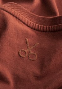 Tessuto color ruggine con una morbida trama, caratterizzato da un design di forbici ricamato in marrone vicino al collo e bordi a coste.