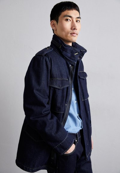 Denim jacket in dark blue with a high collar, zip front, and multiple pockets. Features contrast stitching and a relaxed fit.