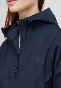 Donna che indossa una giacca blu navy con colletto alto, parzialmente zip, che mostra una camicia bianca sotto.