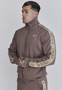 Chaqueta marrón con cremallera y cuello alto, franjas laterales en color crema en contraste con logotipo repetido, y tirador de cremallera de goma.