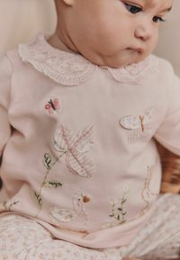 Camisa rosa pálido con cuello de encaje, presenta mariposas bordadas, flores y un pato. Patrón floral en los pantalones a juego. Tela texturizada.