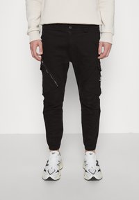 Svarta cargobyxor med avsmalnande ben, utrustade med sidofickor med dragkedja och en logopatch. Tillverkade av ett hållbart material med en slät yta.