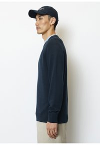 Marineblauer Strickpullover mit gerippten Abschlüssen und Saum, mit einem Rundhalsausschnitt. Getragen über einem hellblauen Hemd, kombiniert mit beigen Hosen und einer schwarzen Mütze.