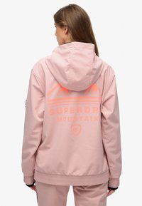 Lichtroze waterdichte jas met capuchon. Heeft "SUPERDRY MOUNTAIN" in oranje opdruk en bergillustraties op de achterkant. Elastische manchetten.