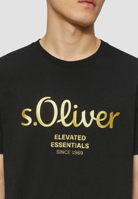 Black cotton T-shirt featuring a gold printed logo that reads "s.Oliver" and text "ELEVATED ESSENTIALS SINCE 1969" beneath it.