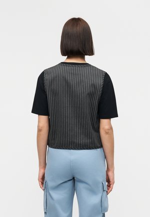 Oseba s kratkimi rjavimi lasmi nosi črno telovnik z drobnimi črtami preko črne kratkoročne srajce in svetlo modre delovne hlače, obračena stran.