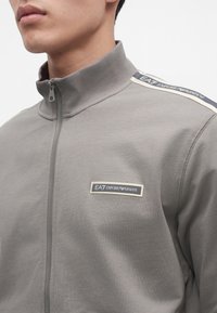 Крупен план на човек, носещ сива яке с цип с логото "EA7 Emporio Armani" на гърдите и ивица на рамото.