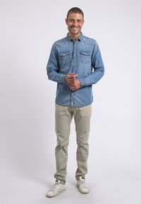 Chemise en denim bleu clair avec deux poches poitrine, col boutonné et manches longues. Associée à un pantalon beige et des baskets blanches.