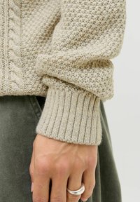 Un pull en tricot beige présentant un corps texturé avec un design en torsades et des poignets côtelés. Main visible, portant une bague en argent.