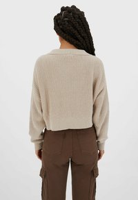 Kvinna med långt, mörkt flätat hår som bär en beige stickad tröja och bruna cargobyxor, sedd bakifrån mot en enfärgad bakgrund.