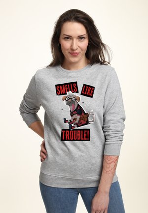 Kobieta w szarej bluzie z kapturem z rysunkowym psem w kostiumie i czerwonym napisem "Smells Like Trouble", stojąca z ręką na biodrze.