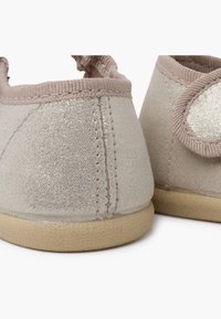 Calzado de color beige claro con una textura suave y brillante, apertura elástica y suela flexible. Presenta un diseño simple y sin costuras.