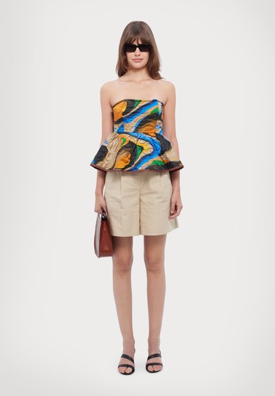 Top bez ramínek s vícebarevným peplum stylem s modrými, oranžovými a černými vzory. Béžové šortky a černé sandály. Modelka drží hnědou tašku.