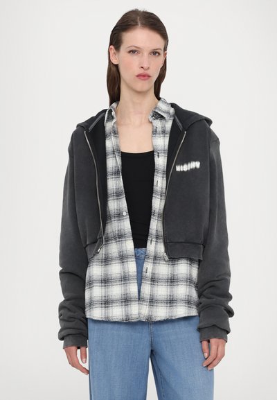 Schwarzer, taillierter Hoodie mit Reißverschluss und grauen Akzenten, getragen über einem schwarzen Tanktop, kombiniert mit einem weiß-schwarz karierten Flanellhemd und blauen Jeans.