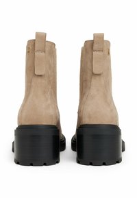 Une paire de bottines en suédine beige avec des talons épais noirs et des tirettes, vues de l'arrière sur fond blanc.