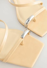 Beige sandaalit, joissa on neliön muotoinen varvasosa, ohuet hihnat ja hopeinen metallinen yksityiskohta varpaiden lähellä, valmistettu sileästä synteettisestä materiaalista.