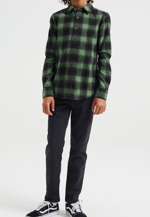 Ein Kind trägt ein langärmliges Hemd in grün-schwarzem Karomuster, schwarze Hosen und schwarz-weiße Turnschuhe und steht vor einem weißen Hintergrund.