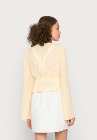 Blusa peplum amarela com flores e mangas longas, apresentando uma cintura elástica e um tecido transparente por cima, combinada com uma saia branca de brocado.