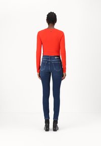 Röd långärmad topp med rund hals, i kombination med åtsittande mörkblå jeans. Outfitten har en logotyp vid halslinningen och bakfickorna.