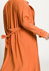 Pomarańczowy płaszcz trencz z paskiem do wiązania, z pojedynczym zapięciem na guzik, długimi rękawami i gładką teksturą materiału.