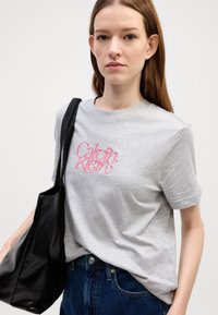 Harmaa puuvillapaita, jossa on lyhyet hihat ja vaaleanpunainen "Calvin Klein" -logo. Mallilla on päällään musta olkalaukkua ja siniset farkut.