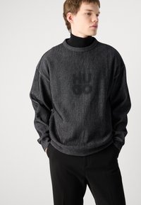 Svart og gråstripet sweatshirt med avslappet passform og rund hals, med subtil logo på baksiden. Brukt med svarte bukser.