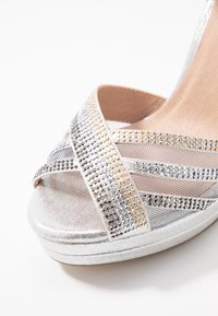 Stříbrné platformové sandály s křížovými metalickými pásky, zdobené flitry ve stříbrné a zlaté barvě, s texturovanou tkaninou a hladkým povrchem.