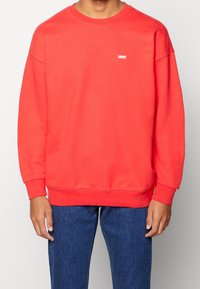 Roter Sweatshirt mit Rundhalsausschnitt und gerippten Bündchen. Verfügt über ein kleines weißes "LEVI'S"-Logo auf der Brust. Kombiniert mit blauen Jeans.
