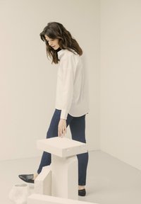 Witte button-up shirt met een relaxte pasvorm, blauwe skinny jeans en zwarte ballerina's. De achtergrond bevat lichtgekleurde geometrische props.