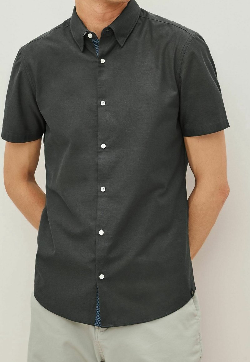Homme portant une chemise gris foncé à manches courtes avec des boutons blancs et un pantalon de couleur claire sur un fond uni.