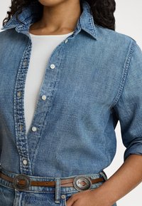 Žena oblečená v modrej džínsovej košeli na gombíky cez biely top, s hnedým koženým opaskom s dvoma striebornými ozdobnými prackami.