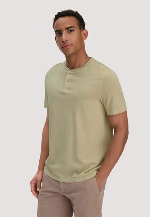 Man met krullend haar die een lichtgroen Henley-shirt met korte mouwen en beige broek draagt, staand met één hand in de zak.