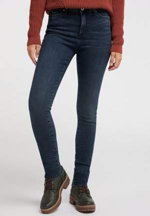 Personne portant un jean skinny bleu foncé, des bottines vert foncé à lacets avec des semelles marron, et un pull en maille couleur rouille.