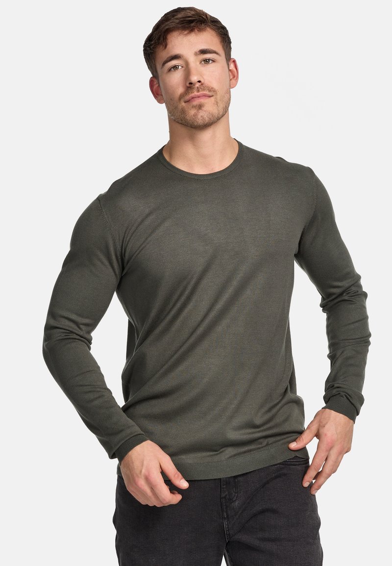 Pull à manches longues vert foncé avec un col rond, des poignets côtelés et un tissu texturé, porté sur un jean noir.