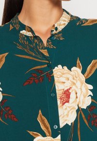 Blusa floreale in tessuto teal con fiori color crema e oro e accenti rossi. Il collo rotondo e i piccoli bottoni valorizzano il design.