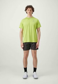 Man standing wearing lime green short-sleeve shirt, black shorts, black socks, and white running shoes against plain background.