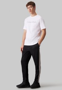 T-shirt bianca in cotone con logo Calvin Klein; pantaloni sportivi neri con strisce laterali e logo; sneakers nere. Design semplice e moderno.
