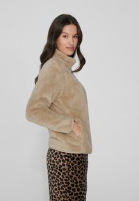 Sudadera de forro polar beige con cuello alto, mangas largas y bolsillos laterales. Combinada con pantalones de leopardo marrón. Tejido suave y texturizado.