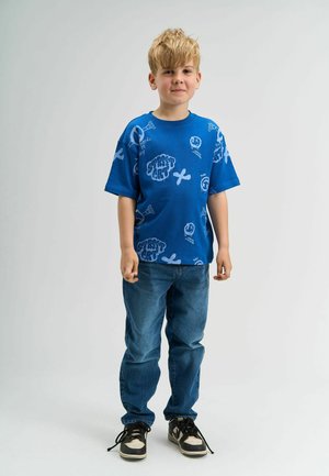 Tricou grafic albastru cu design de artă stradală, mâneci scurte, asociat cu blugi blue faded și adidași negri cu accente albe.