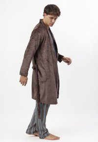 Joven en una bata de baño de felpa marrón y pantalones de pijama de rayas azules, de pie descalzo sobre un fondo blanco, mirando hacia abajo con los brazos ligeramente extendidos.