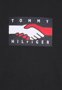 Černé tričko s grafickým motivem dvou rukou, které si podávají ruce, jedna bílá a jedna červená, s textem "TOMMY HILFIGER" pod nimi v bílé barvě na modrém pozadí.