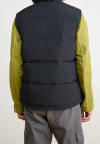 Kamizelka ocieplana z czarną górną częścią i oliwkowozielonymi rękawami. Gładka faktura z widocznymi przeszyciami i wysokim kołnierzem.