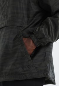 Giacca a vento verde scuro con un motivo camouflage testurizzato, polsini elasticizzati e tasca frontale con apertura superiore.