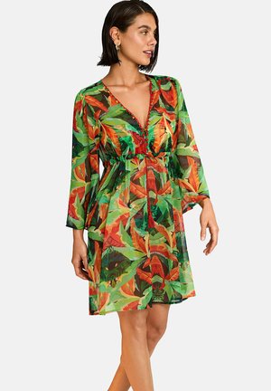 Femme portant une robe mi-longue à motifs de feuilles vertes et orange, col en V profond et manches trois-quarts, regardant sur le côté.