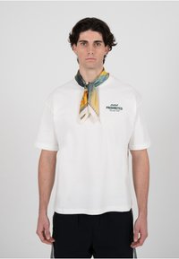 Hvit bomulls-T-skjorte med korte ermer, med en blå "PROHIBERTE"-logo. Et fargerikt silkesjal knyttet rundt halsen gir en kontrasterende detalj.