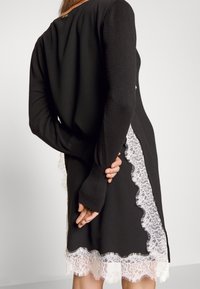 Femme portant une robe noire à manches longues avec une bordure en dentelle blanche sur les côtés et l'ourlet, mains jointes derrière le dos, debout devant un fond uni.